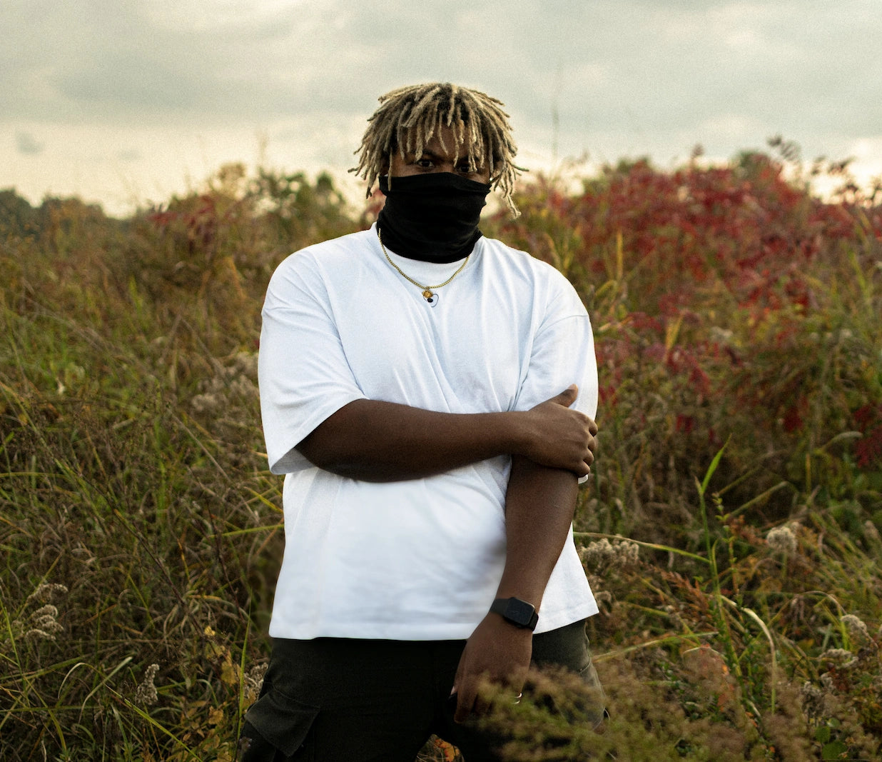 Person wearing a white shirt and black mask standing in a field with tall grass and red flowers.